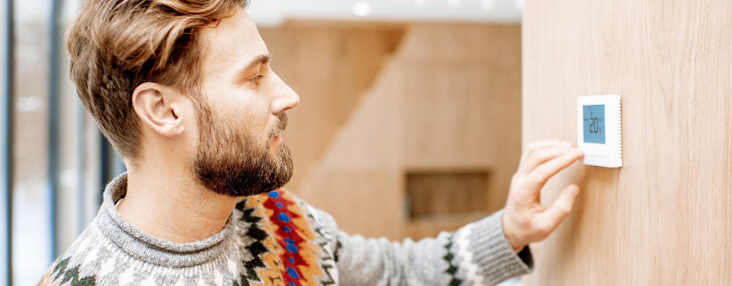 Ein Mann mit Bart bedient ein digitales Thermostat, um die Heizungssteuerung im Raum anzupassen. Die Anzeige des Geräts zeigt eine Temperatur von 20°C. Die Szene spielt in einem hellen, modernen Zuhause mit Holzelementen.