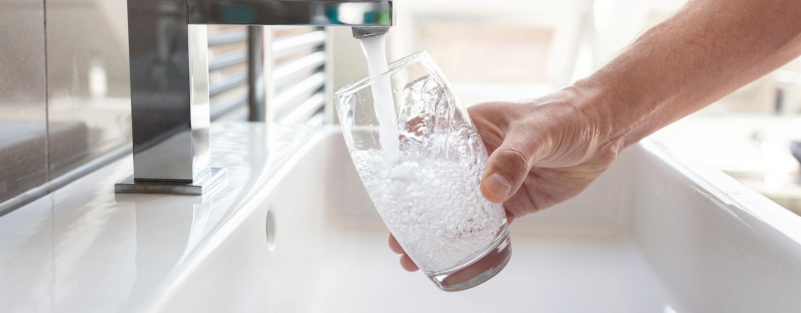 Ein Wasserglas wird von einer Hand unter den Hahn gehalten, während frisches Trinkwasser mit sichtbaren Blasen eingefüllt wird.