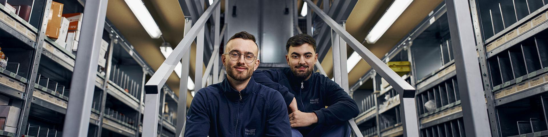 Zwei junge Männer in Arbeitskleidung von bad und heizung sitzen auf einer Metalltreppe in einem Lagerraum, umgeben von Regalen voller Ersatzteile und Verpackungen. 