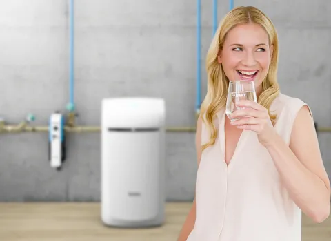 Lächelnde Frau mit einem Glas hochwertigem Trinkwasser in der Hand, im Hintergrund ist eine technische Wasseraufbereitungsanlage sichtbar.