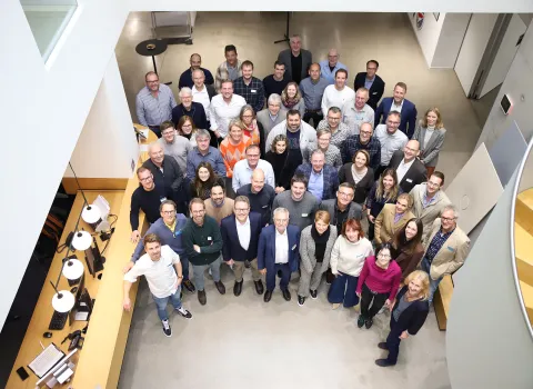 Eine große Gruppe von Menschen steht dicht beieinander in einem modernen Bürogebäude und blickt lächelnd nach oben in die Kamera.