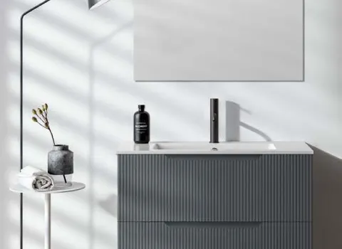 Modernes Badmöbel mit geriffelter Front in Grau unter quadratischem Spiegel und seitlicher Stehlampe.