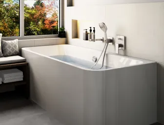 Wandstehende Badewanne mit glänzender Oberfläche und Wandarmatur. Links steht eine Bank mit Kissen und Handtüchern, dahinter ein großes Fenster mit Herbstblick.