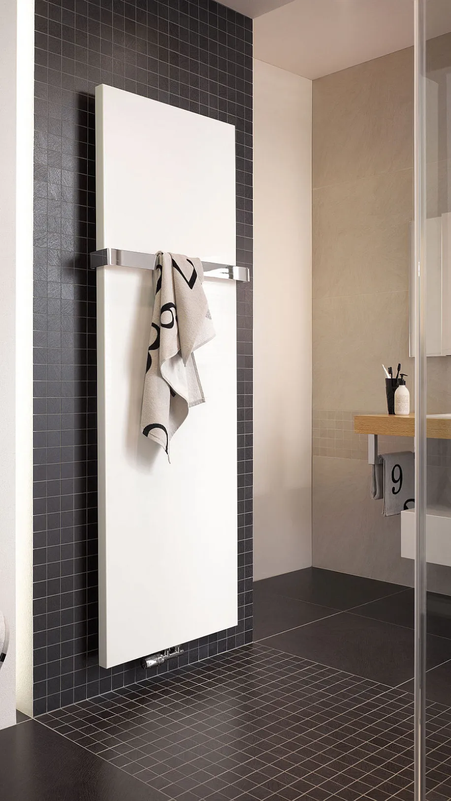 Ein weißer Designheizkörper von HSK ist an einer Wand mit dunklen Fliesen montiert, daran hängt ein helles Handtuch mit schwarzen Zahlen.