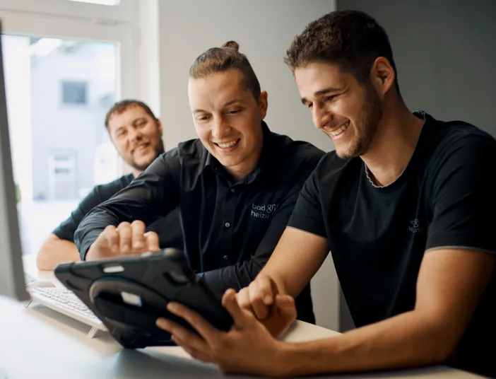 Drei Kollegen stehen an einem Schreibtisch und arbeiten gemeinsam an einem Tablet, das von einem der Männer gehalten wird. 