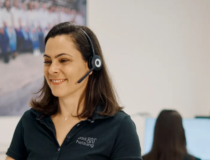 Eine Frau mit einem Headset sitzt lächelnd an einem Schreibtisch in einem modernen Büro.