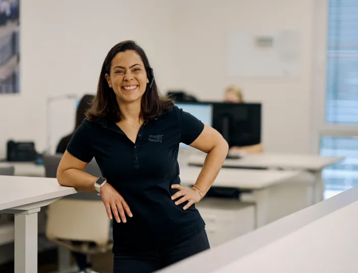 Eine lächelnde Frau steht in einem modernen Büro, leicht an einen Tisch gelehnt. Sie trägt ein Headset und ein dunkelblaues Poloshirt mit einem Firmenlogo.