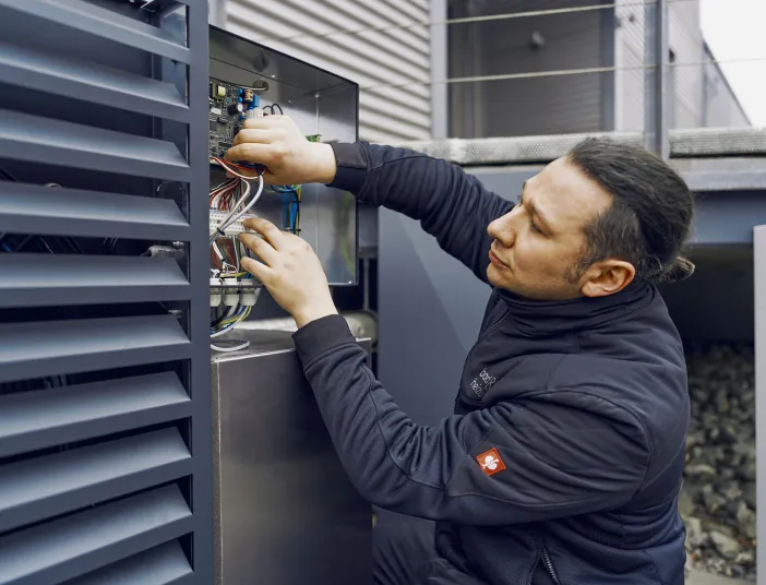 Ein Techniker überprüft eine Elektroinstallation an einer Außeneinheit, während er sorgfältig Kabel in einem offenen Schaltkasten anschließt.