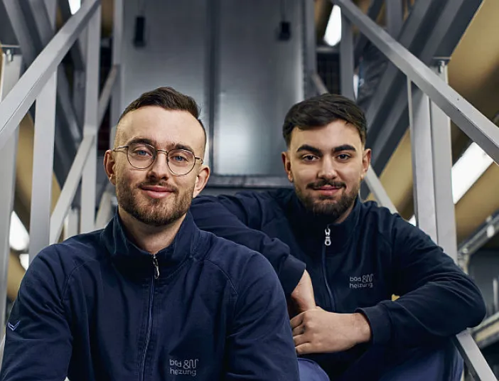 Zwei junge Männer in Arbeitskleidung von bad und heizung sitzen auf einer Metalltreppe in einem Lagerraum, umgeben von Regalen voller Ersatzteile und Verpackungen. 