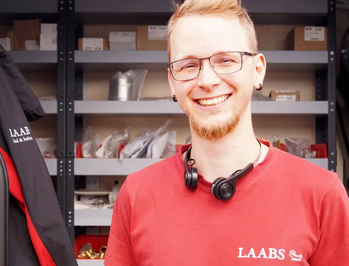 Ein junger Kundendienstmonteur SHK der Firma Laabs mit Brille und Kopfhörern um den Hals lächelt in die Kamera.