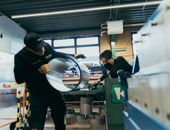 Zwei Auszubildende stehen in einer modernen Werkstatt und arbeiten konzentriert an einem runden Metallstück.