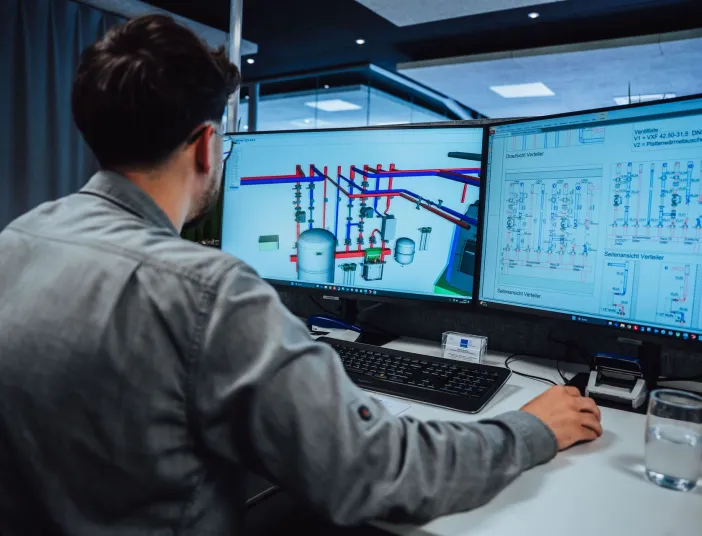 Ein technischer Systemplaner blickt auf zwei Monitore, die komplexe technische Zeichnungen und ein 3D-Modell einer Rohrleitungsinstallation anzeigen.