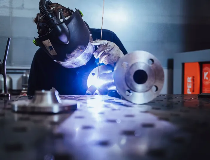 In einer professionellen Werkstatt schweißt ein Arbeiter unter einem Schutzhelm ein rundes Metallbauteil.